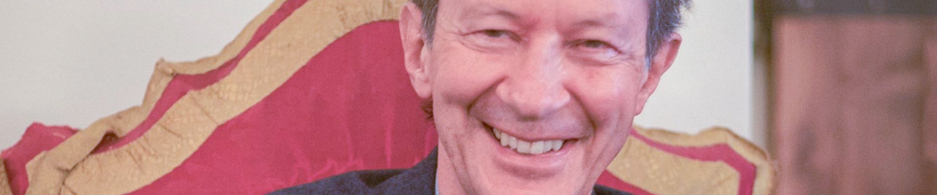 Giorgio Agamben siede sorridente su una sedia ornata, indossando una giacca elegante sopra una camicia chiara e un maglione scuro, con un libro sfocato in primo piano.