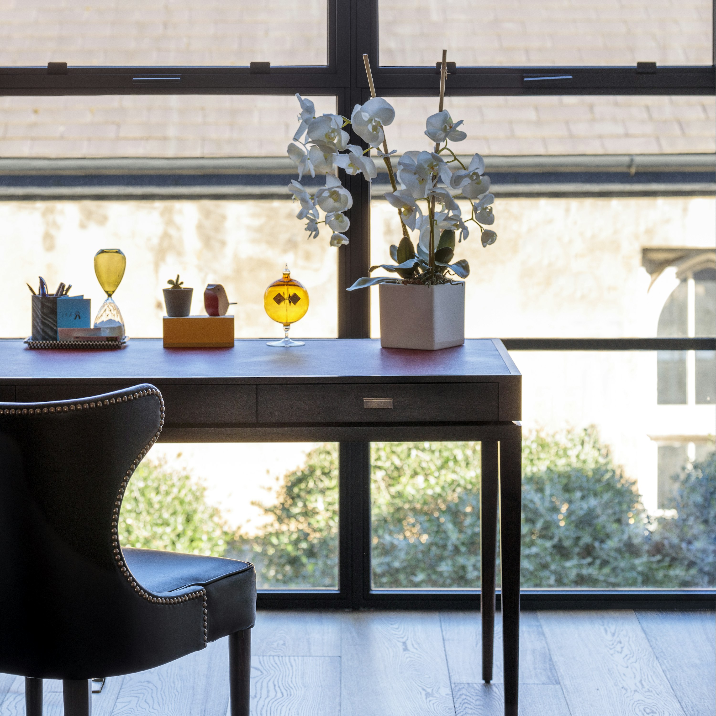 A writing desk with a leather top and a wingback chair in front of large windows in a home study, framing garden views.