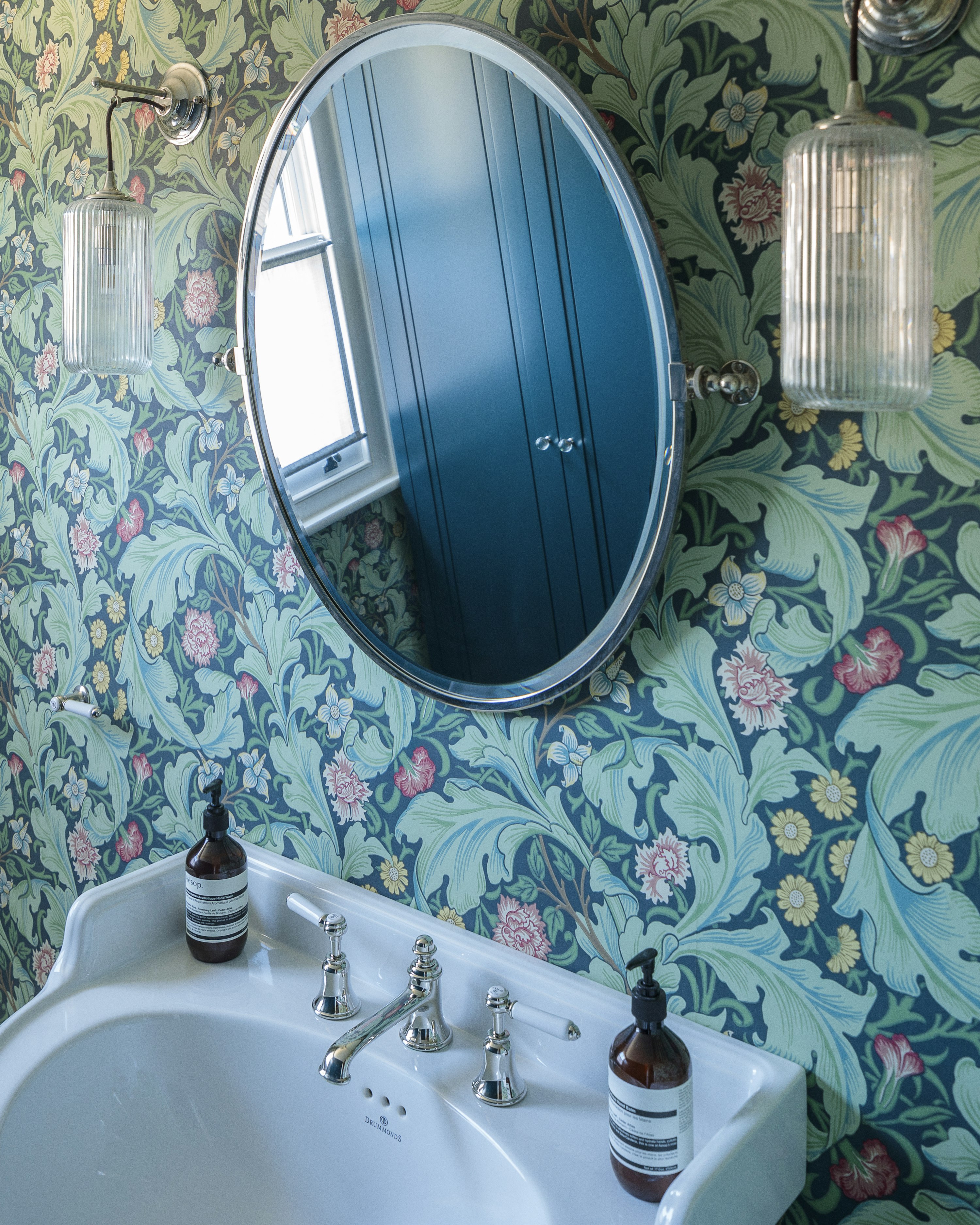 A small WC with a traditional porcelain sink, oval mirror and fluted sconces, on colourful, botanical William Morris wallpaper.