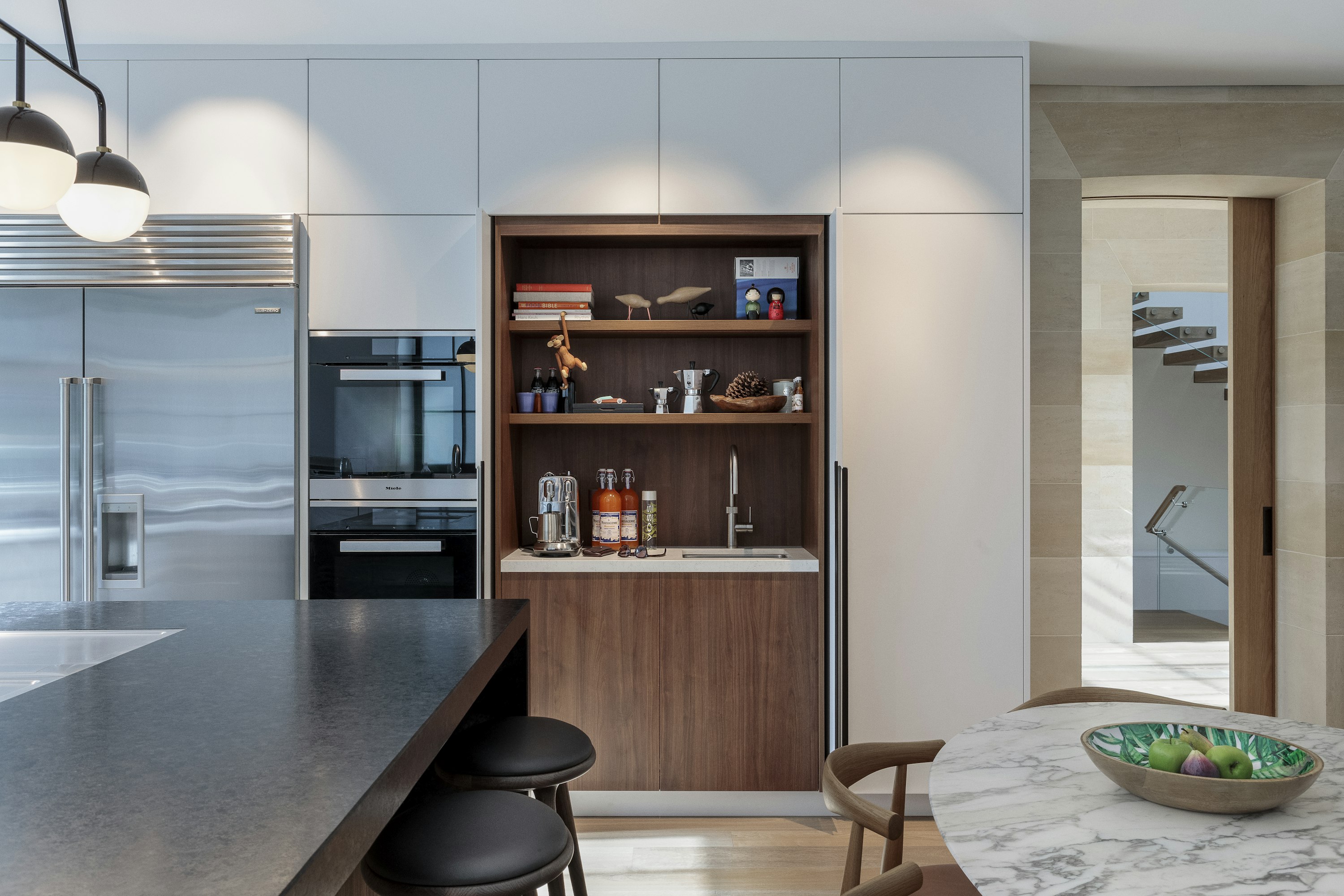 Minimalist kitchen with white cabinets and stone kitchen island. A built-in wooden coffee bar is next to an integrated fridge.