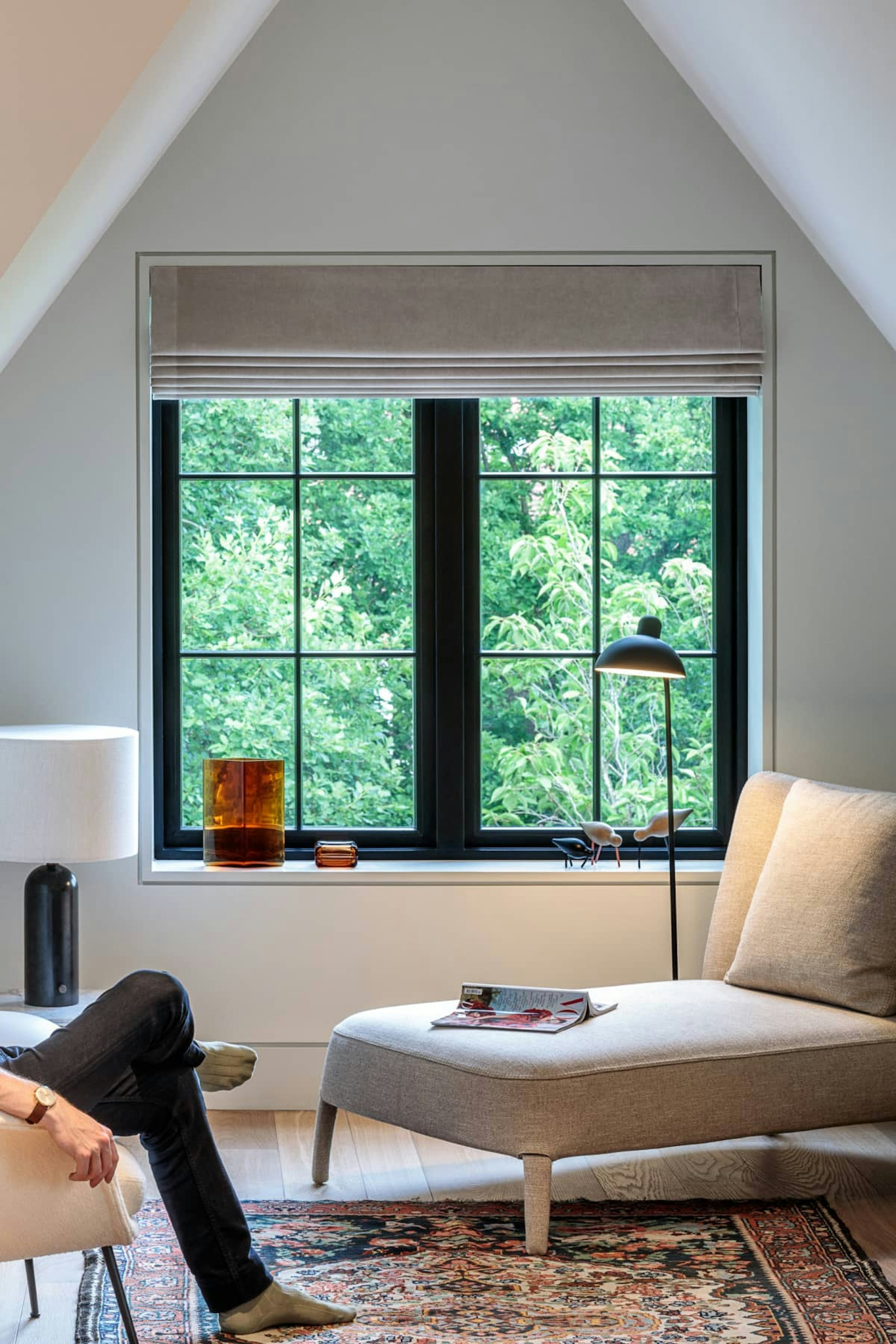A reading corner in a loft bedroom with neutral interiors. A man is sitting next to a chaise lounge, looking into the garden.