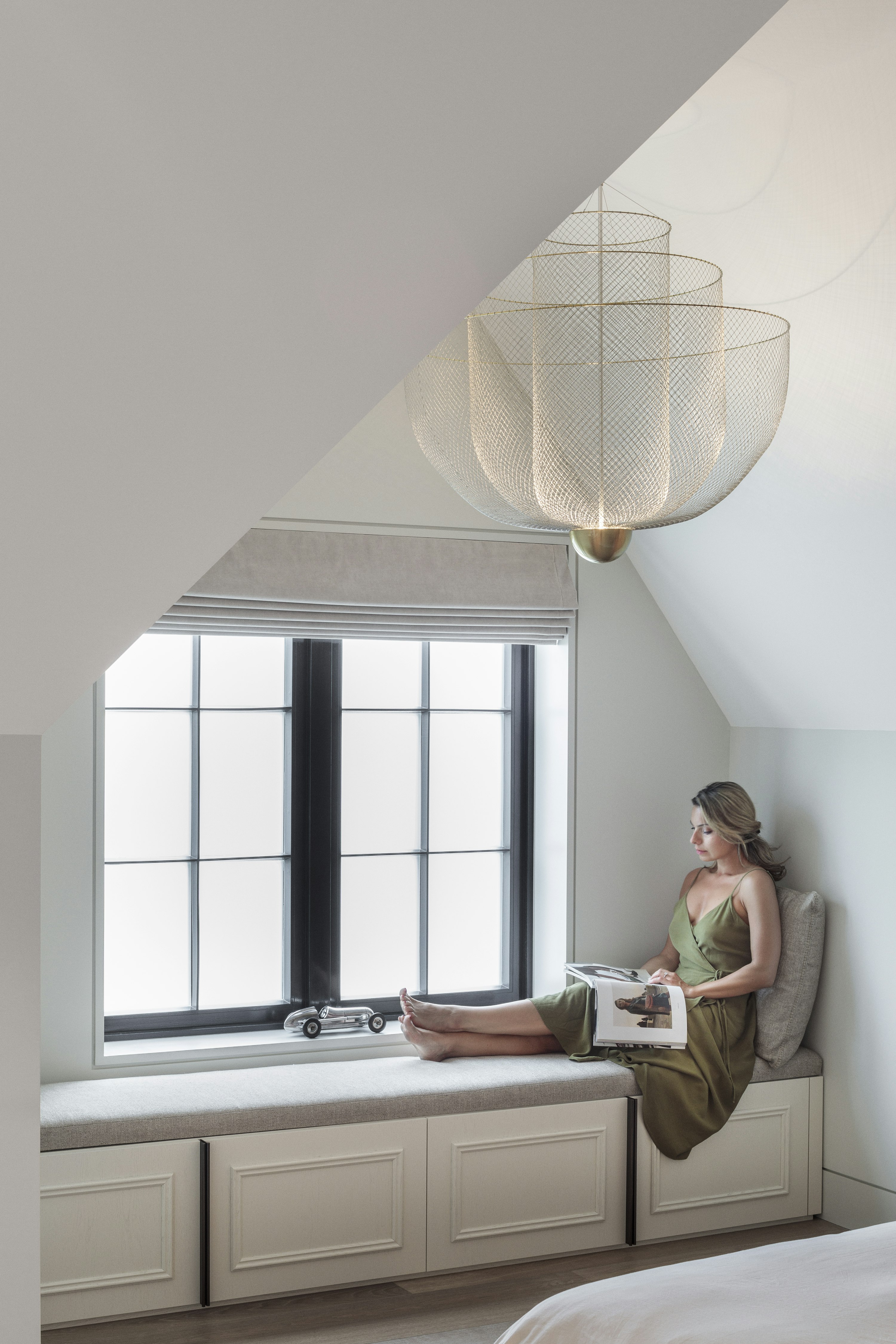 A woman reads a magazine on a window seat with built-in storage, in a light bedroom with a filigree wire chandelier above.