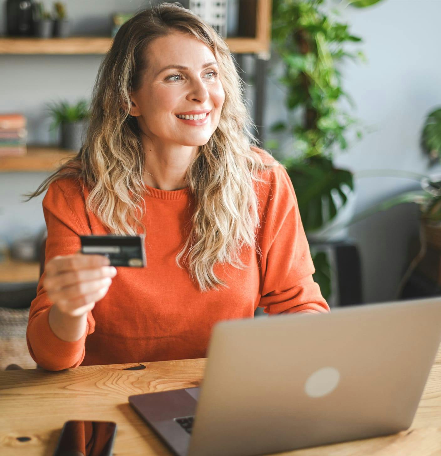 Woman in an orange top holding a credit card in front of a laptop