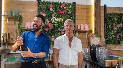 Rylan and Lee stand behind a bar. Rylan is holding a bottle of prosecco