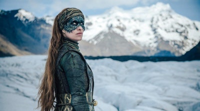Woman with long hair and black mask stands in front of snowy mountain range