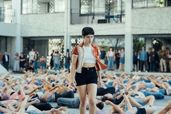 A young woman stands amongst a group of people  lying on the floor around her