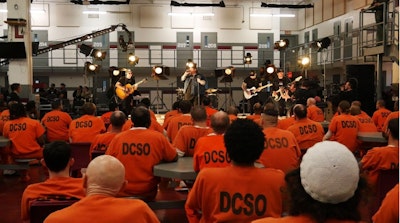 A band performs onstage for a large group of prisoners clad in orange jumpsuits