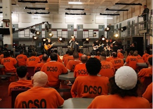 A band performs onstage for a large group of prisoners clad in orange jumpsuits