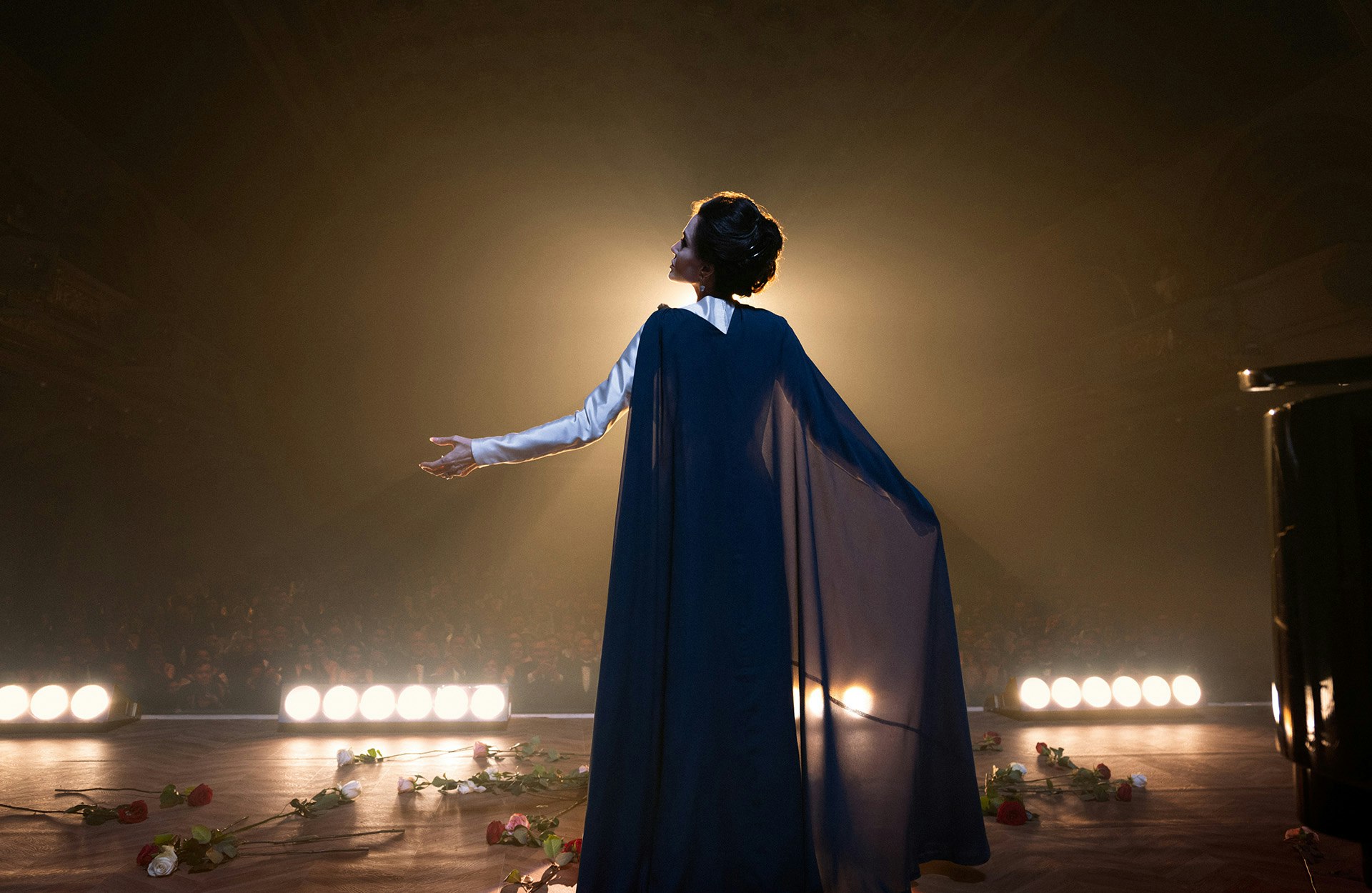 Angelina Jolie stars as Maria Callas in Pablo Larrain's MARIA A woman stands on a stage, whose floor is strewn with roses. The woman, wearing a long blue cape, stands with her arms spread wide. A spotlight shines on her creating a halo effect around her silhouette.