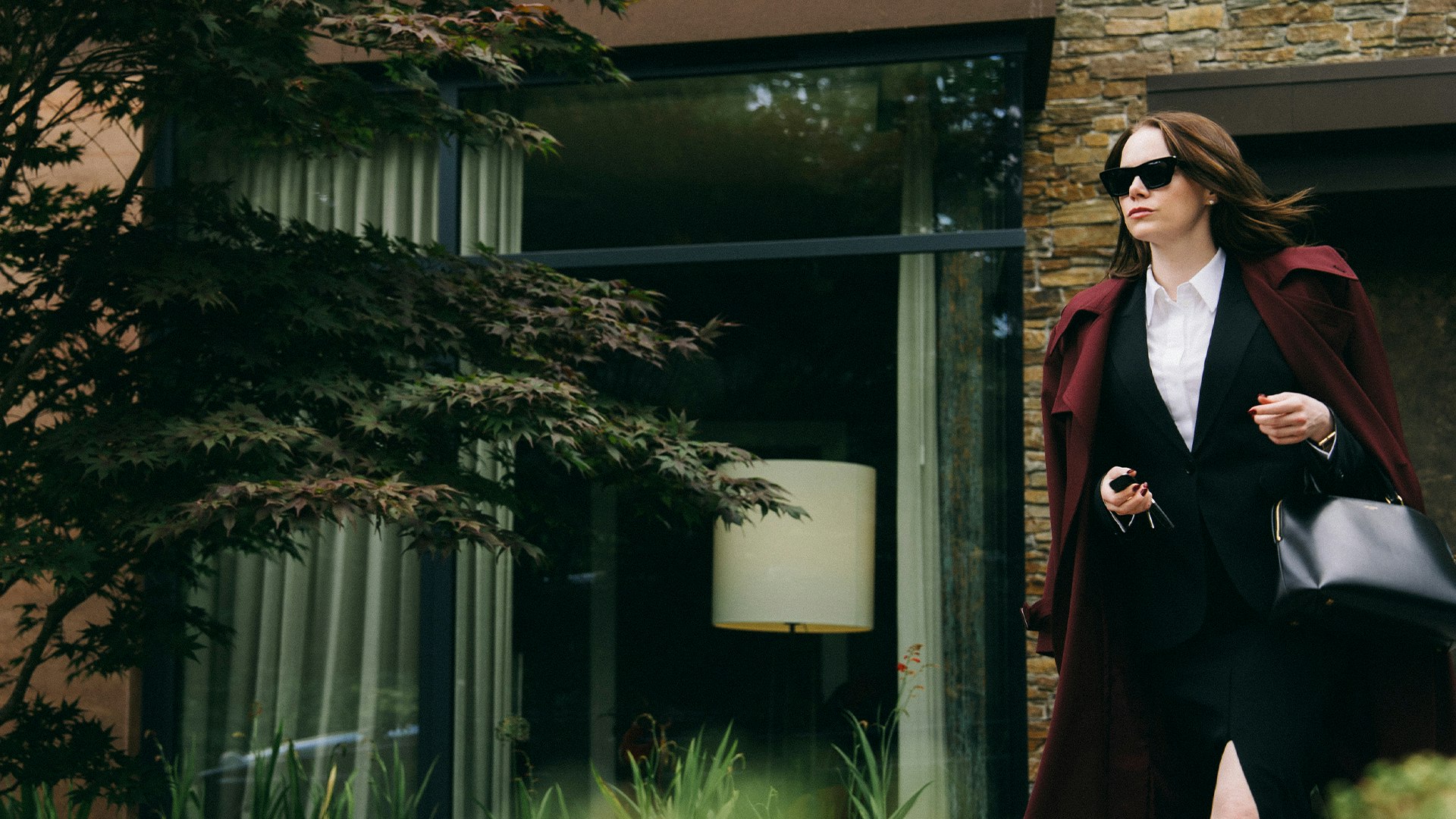 Emma Stone stars in Yorgos Lanthimos' latest cinematic epic, Bugonia A woman dressed in a black skirt suit with a burgundy coat draped over her shoulders and wearing black sunglasses, walks outside a brick-fronted wall with a giant glass-paned window