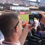 Image of a man using his mobile phone screen to view through his binoculars at a sports game
