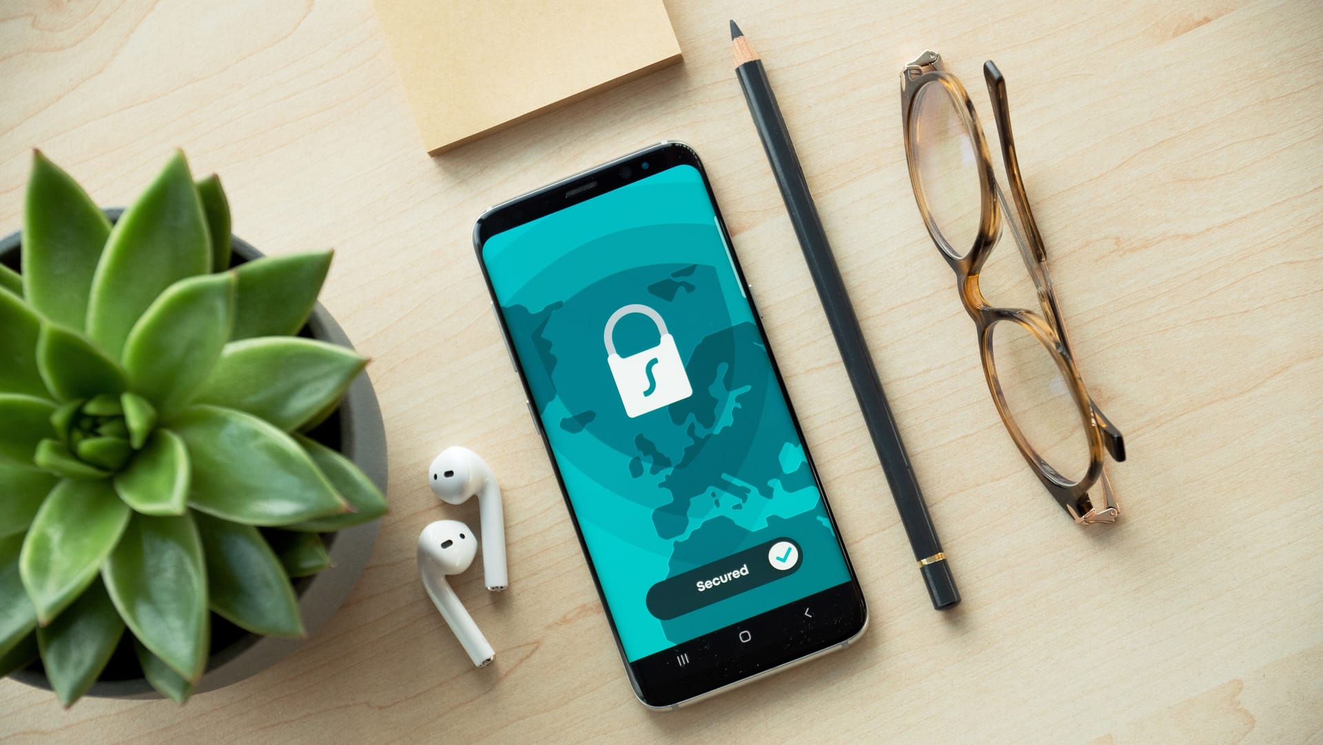 A smartphone with a lock on the screen lies on a table next to a pair of earbuds, a pencil, glasses and a plant.
