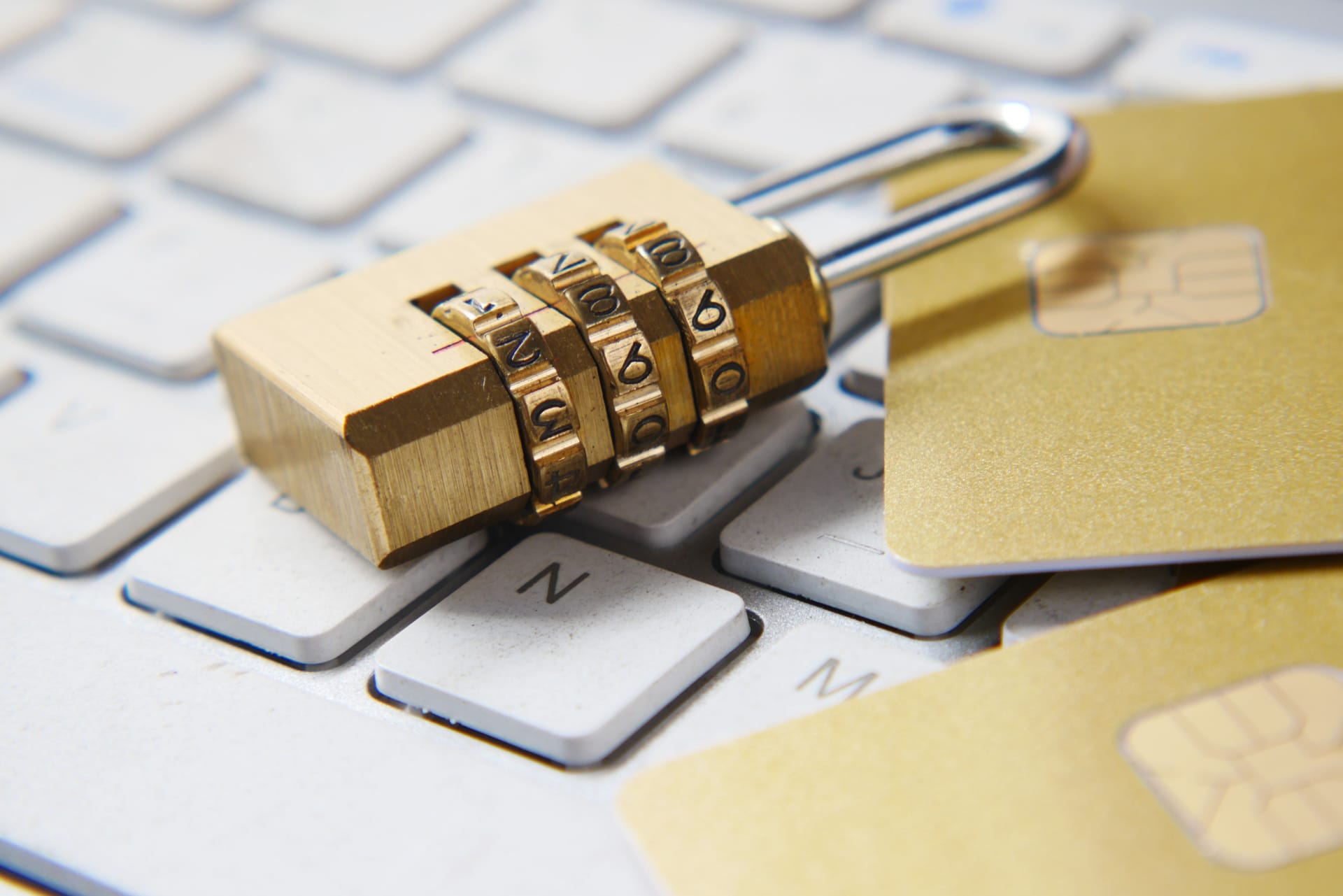 A golden number padlock placed on a white keyboard with two gold credit cards on the right, slightly out of shot.