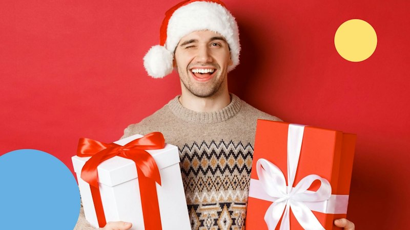Man in sand beige, crew neck sweater with Christmas pattern holding two presents. One present has red wrapping the other white. The man is smiling and winking while wearing a Santa hat, set against a red background.