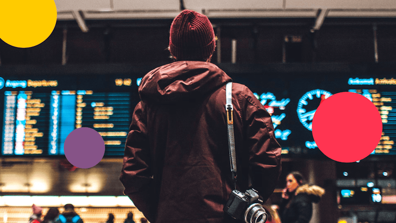 Man in a maroon jacket and fisherman hat with a camera hanging on his shoulder, looks away from the camera shot while looking at the departures board.