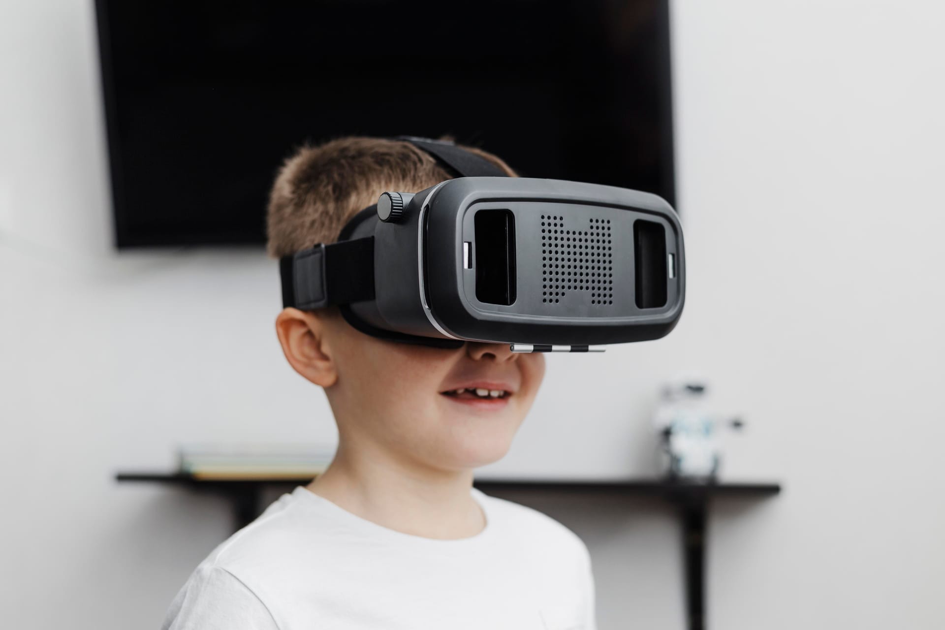 Ein Junge vor einer weißen Wand mit einem Fernseher, der lächelt und eine Virtual Reality-Brille trägt.
