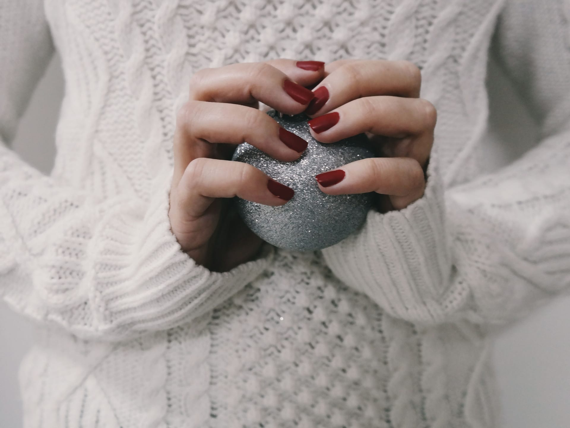 Woman’s hand with red nails holding a silver Christmas bauble with a white sweater