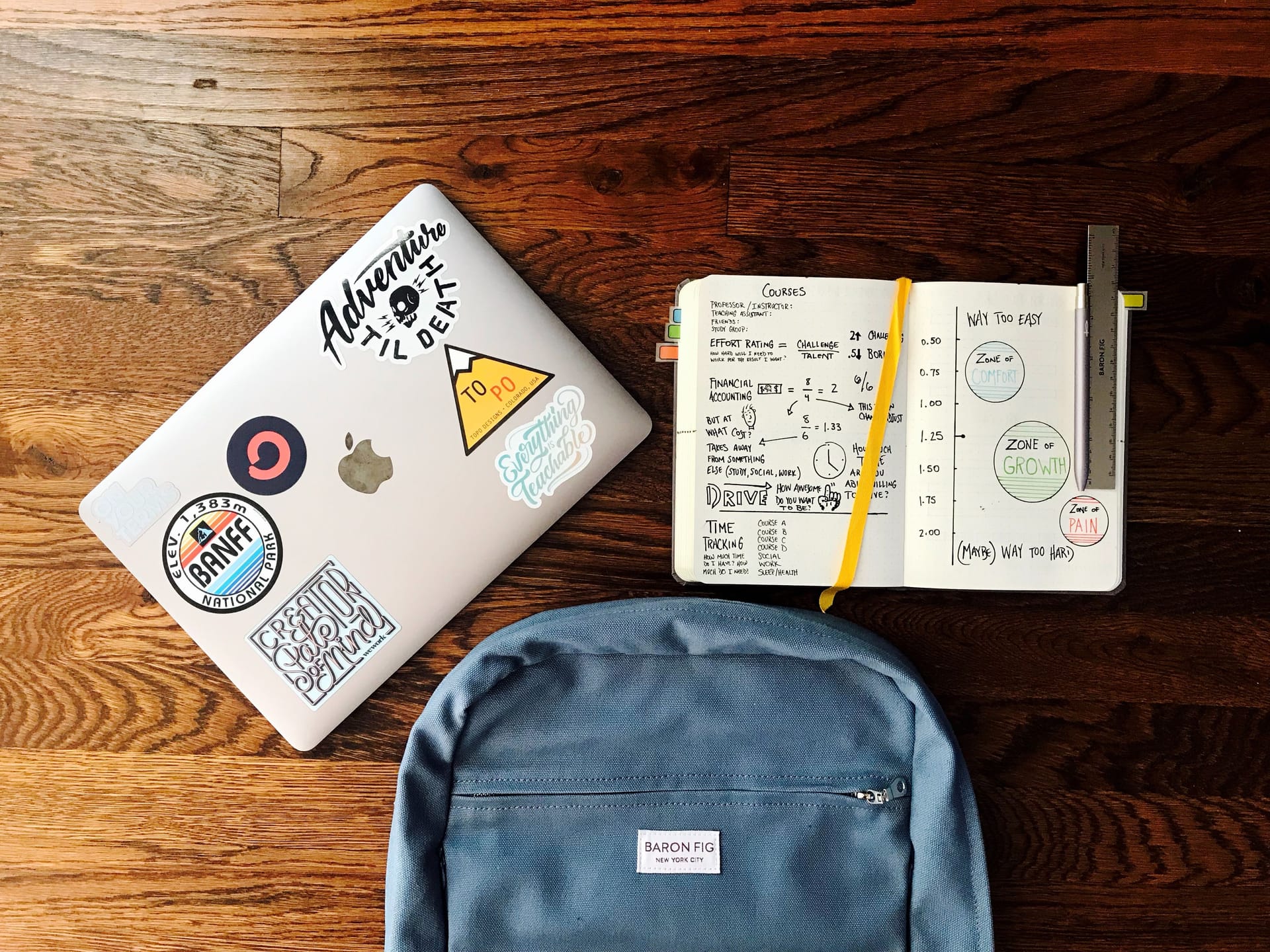 A macbook, notebook and backpack against a wooden table background.