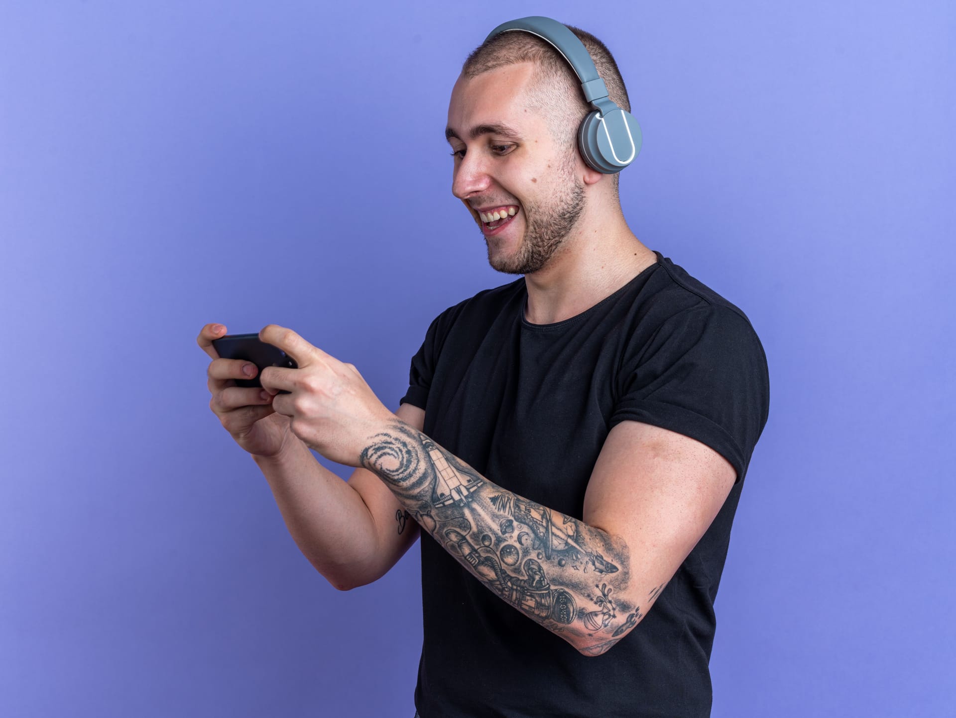 A happy-face man with a headset holding a mobile phone with 2 hands, wearing tattoos and a black t-shirt