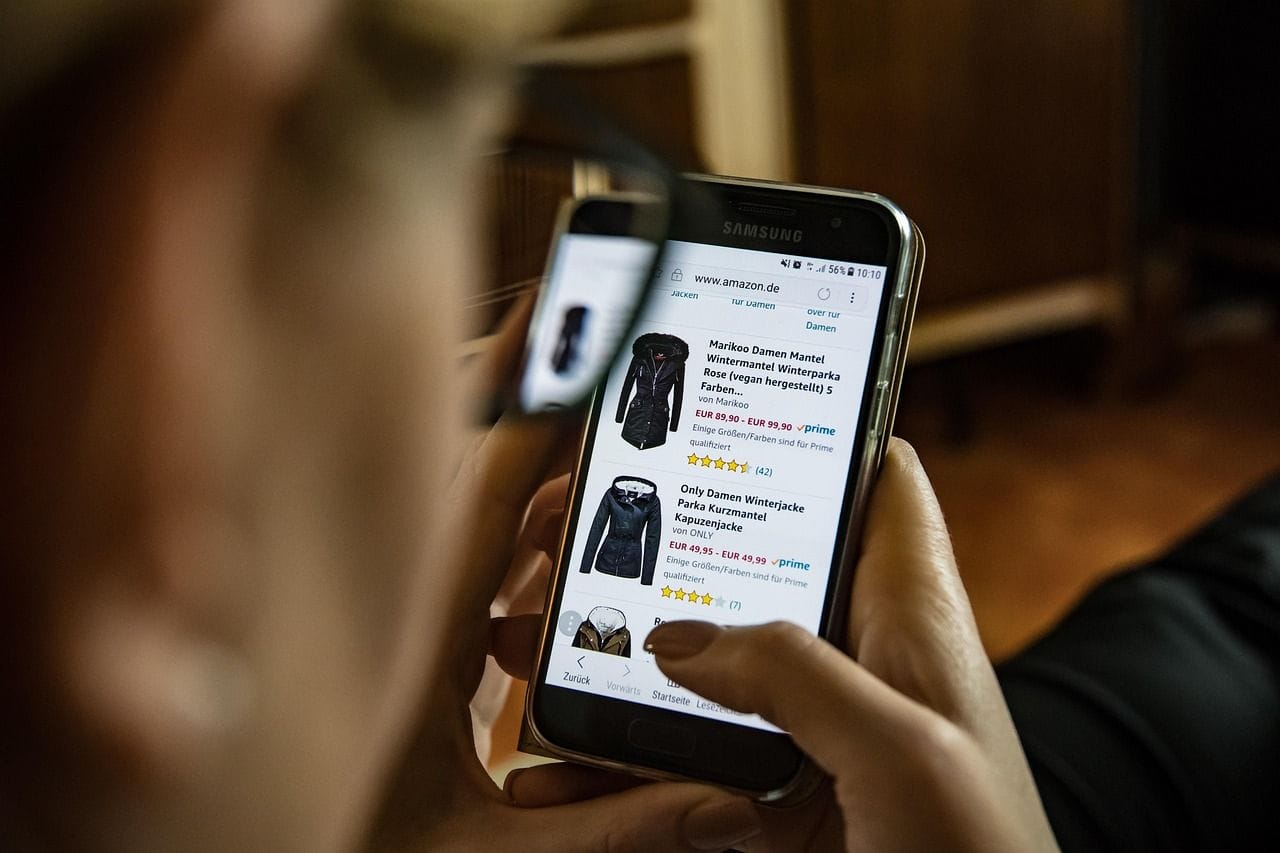 Kobieta robi zakupy online na smartfonie, przeglądając ofertę kurtek w sklepie internetowym.