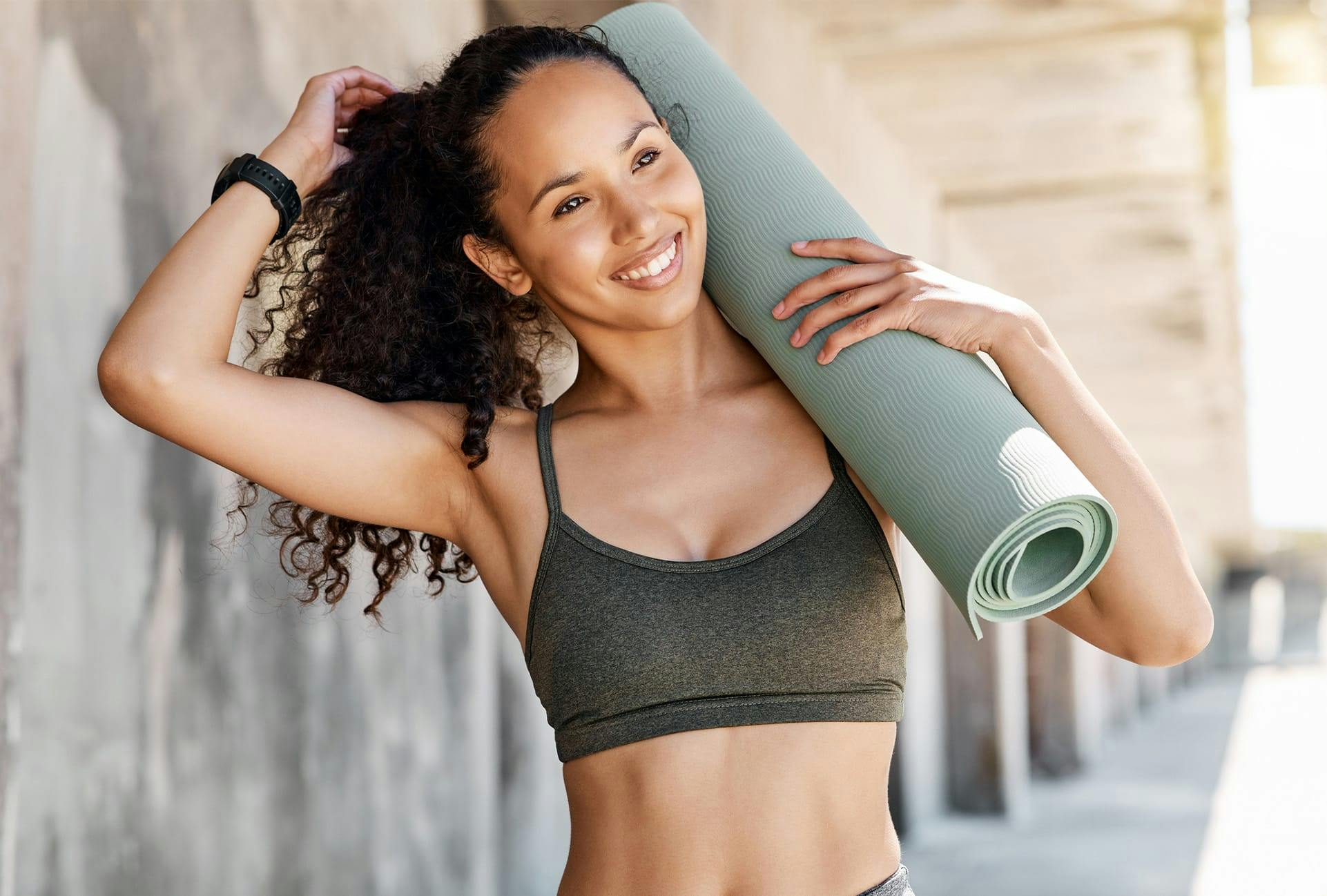 smiling woman with curly hair wearing athletic wear holding yoga mat