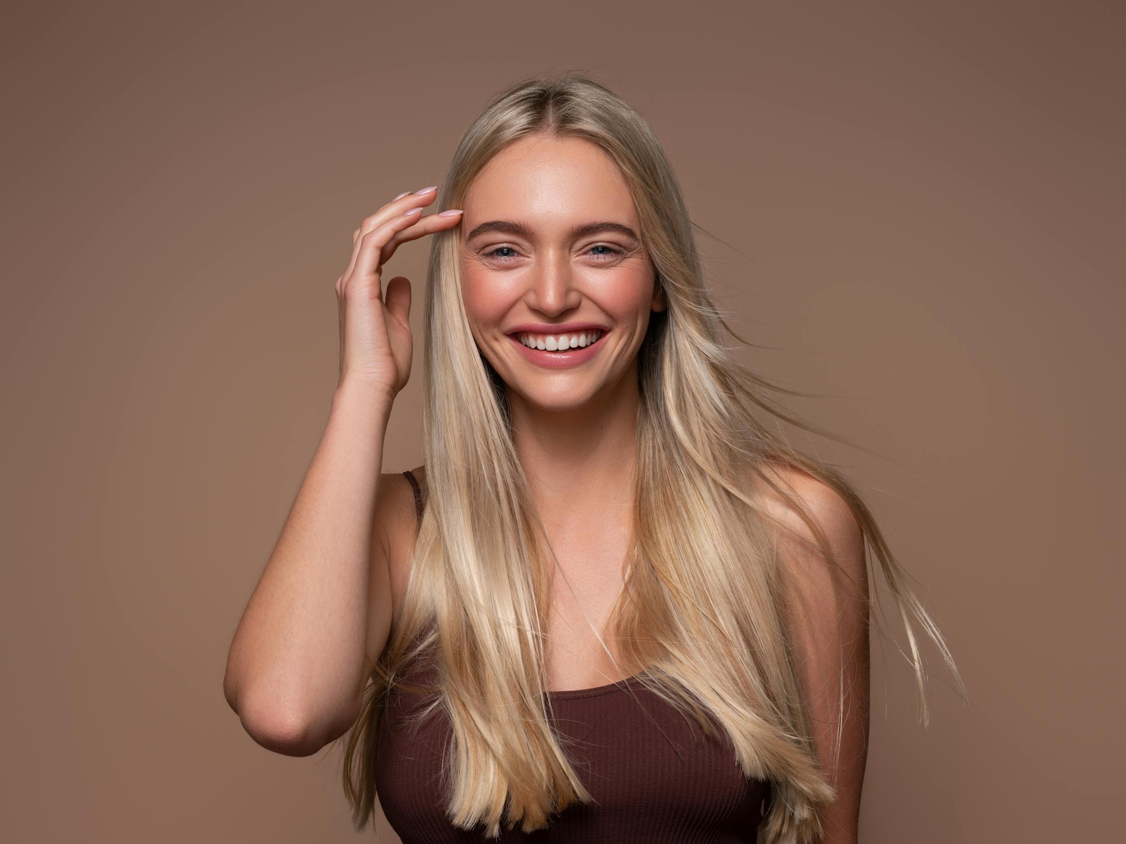 A young woman with long blonde hair is smiling as she has her portrait taken. One hand is up by her forehead. She has beautiful, wrinkle free skin to emulate what the champagne cocktail of botox and filler could achieve.