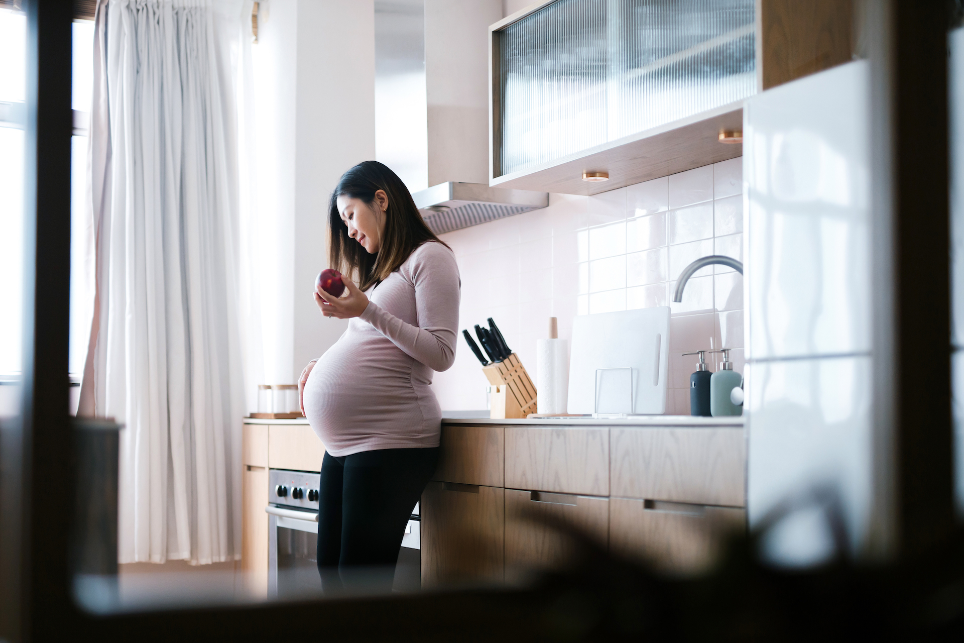 Smiling young Asian, pregnant woman touching her belly, beautiful modern kitchen