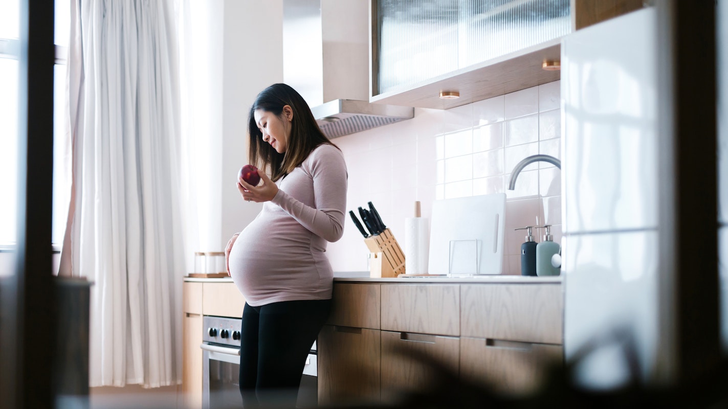 Smiling young Asian, pregnant woman touching her belly, beautiful modern kitchen