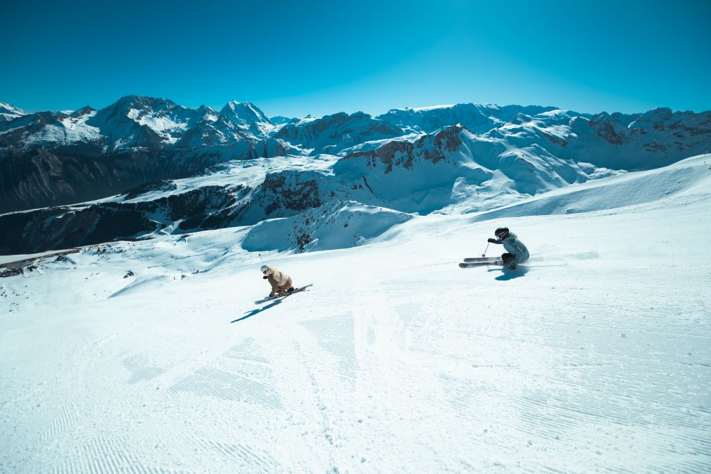 2 skieurs descendent une piste damée, face à eux une chaine de montagne enneigée sous un ciel bleu