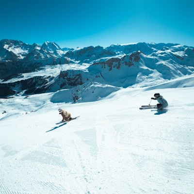 2 skieurs descendent une piste damée, face à eux une chaine de montagne enneigée sous un ciel bleu