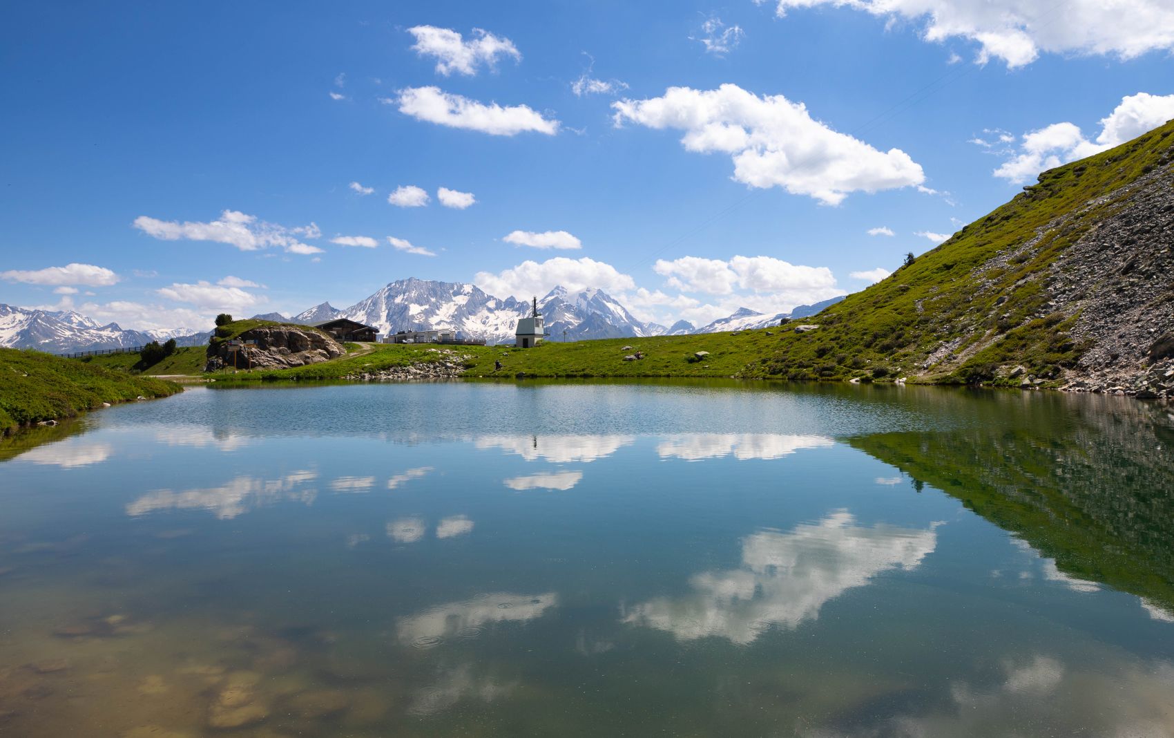 Un lac alpin calme reflète des nuages et des sommets enneigés sous un ciel bleu.