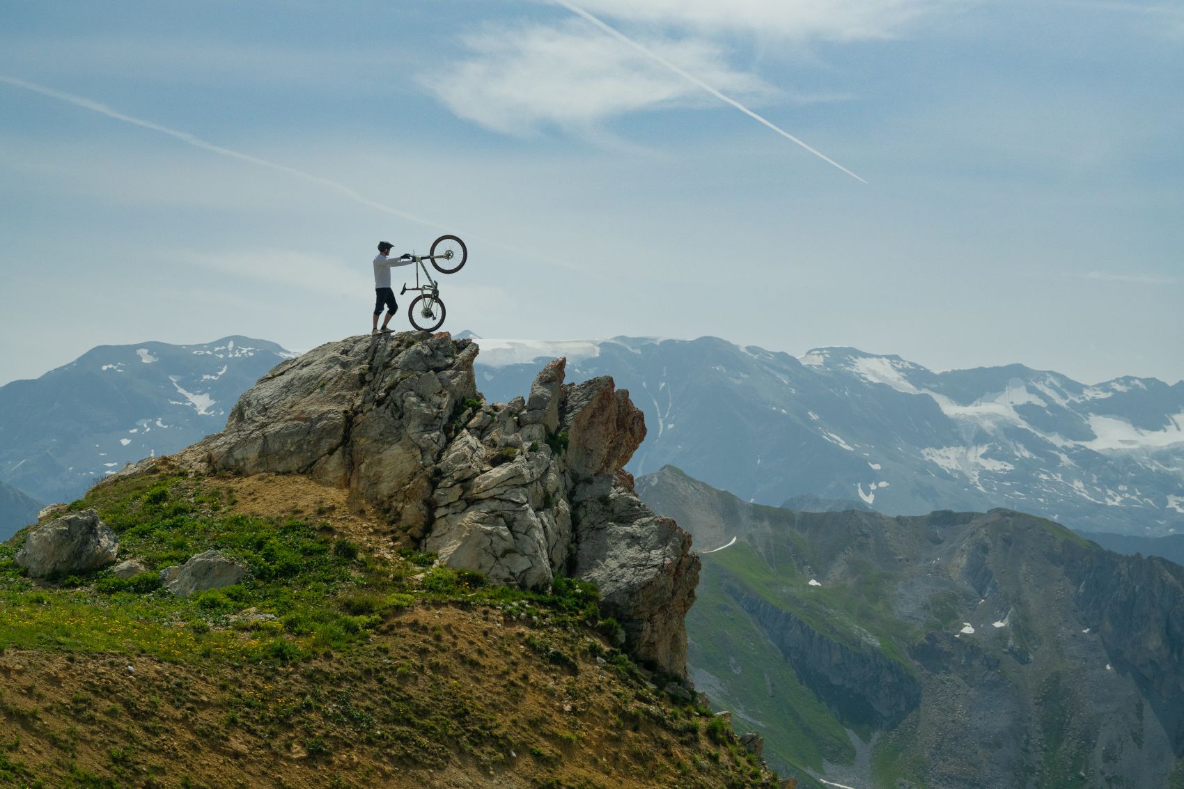 Un cycliste debout avec son vélo au sommet d’un rocher, face aux montagnes enneigées.