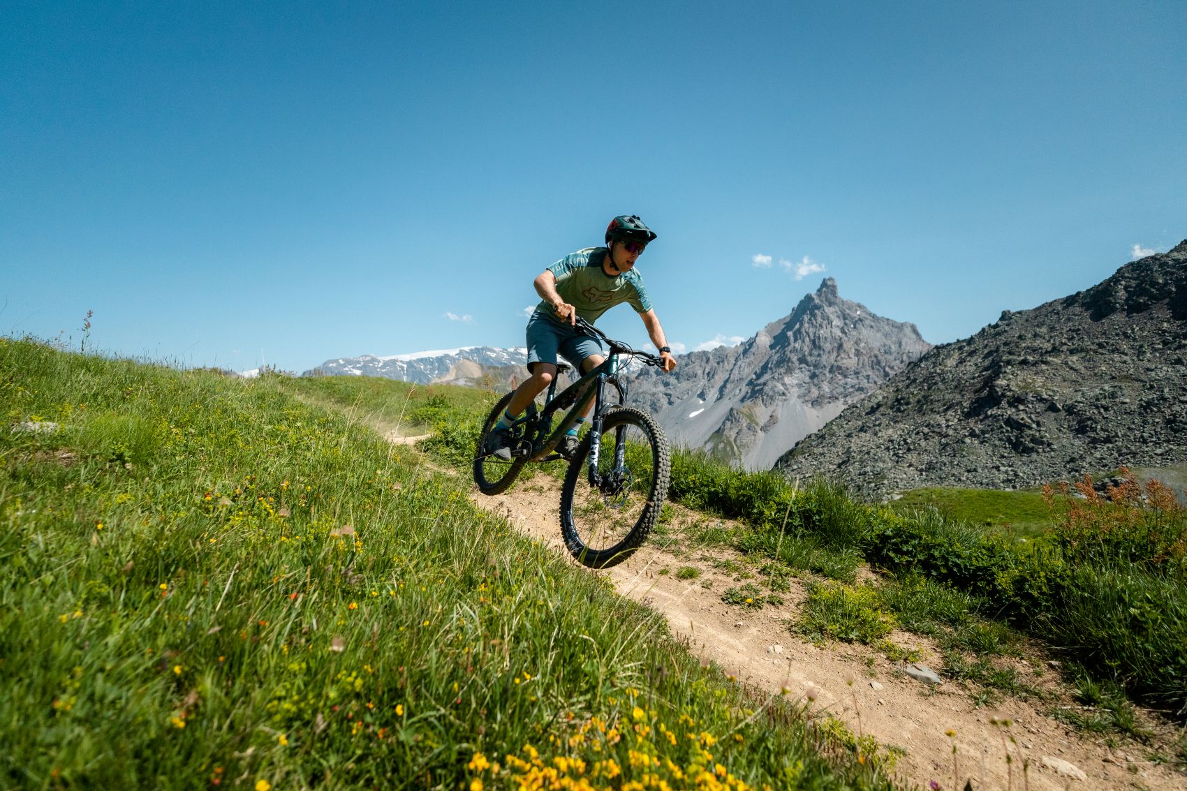 A cyclist is riding downhill on a narrow trail, with mountains in the background