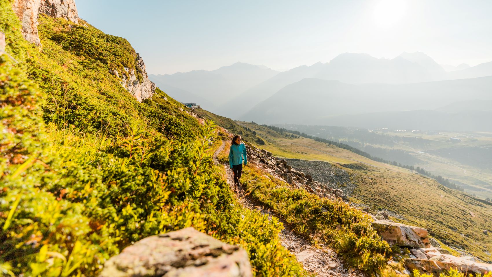 Une randonneuse marche sur un sentier de montagne, entourée de végétation et de sommets en arrière-plan.