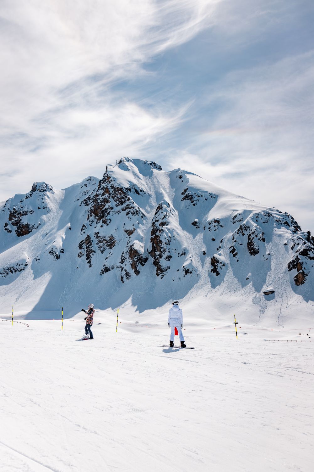 Two snowboarders are riding down a snow-covered slope at the foot of an imposing mountain covered in snow and rocks, beneath a blue sky dotted with wispy clouds.