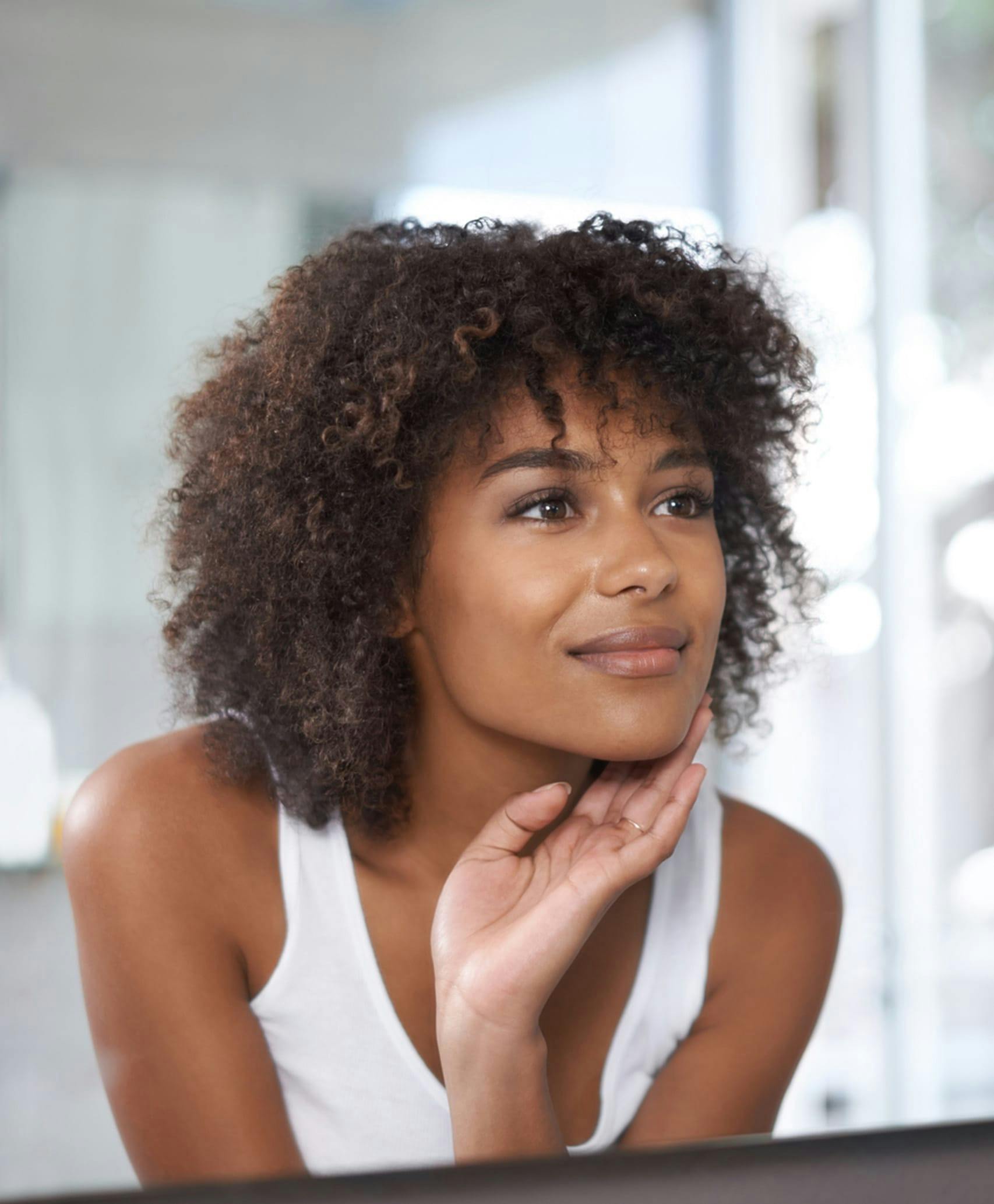 Woman in white tank top looking in mirror with hand on chin.