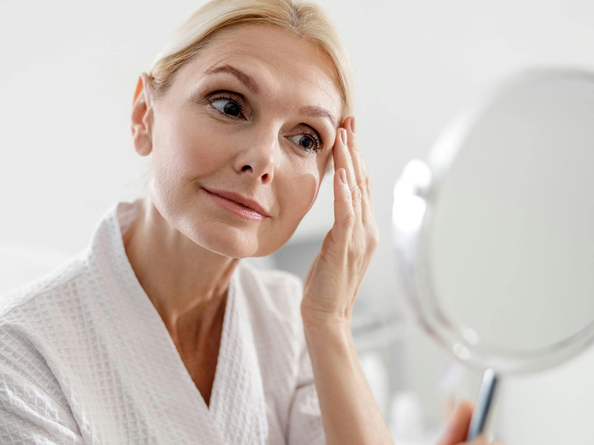 Woman in bathrobe holding mirror looking at face.