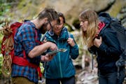 Ein Mann und zwei Frauen schauen auf eine Karte, sie stehen in einem Wald, tragen Rucksäcke und Outdoor-Bekleidung.