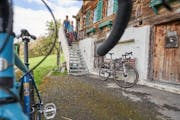 Im Vordergrund ein Velo in der Unschärfe, im Hintergrudn eine Hütte, zwei Personen kommen eine Treppe hinunter.