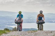 Zwei Personen fahren mit dem Gravelvelo einen Hang hinauf, an den Lenkern ist eine Lenkertasche befestigt, im Hintergrund sieht man einen See.