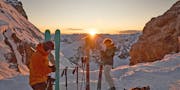 Zwei Personen mit Tourenski mit Sonnenaufgang im Schnee.