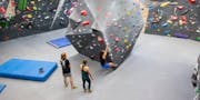 Drei Personen in einer Boulderhalle, jemand bouldert, die anderen schauen zu.
