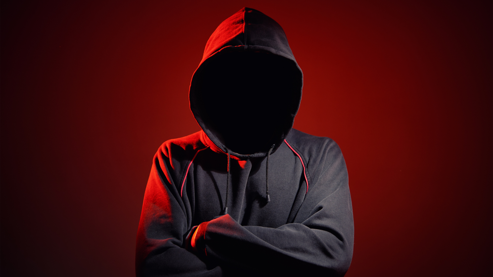 A mysterious figure in a hooded sweatshirt with a faceless void, illuminated by red lighting, symbolizing a cyber threat or hacker persona.