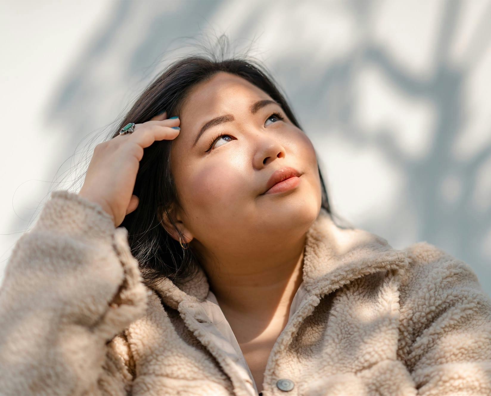 woman in fluffy jacket looking up with hand on temple