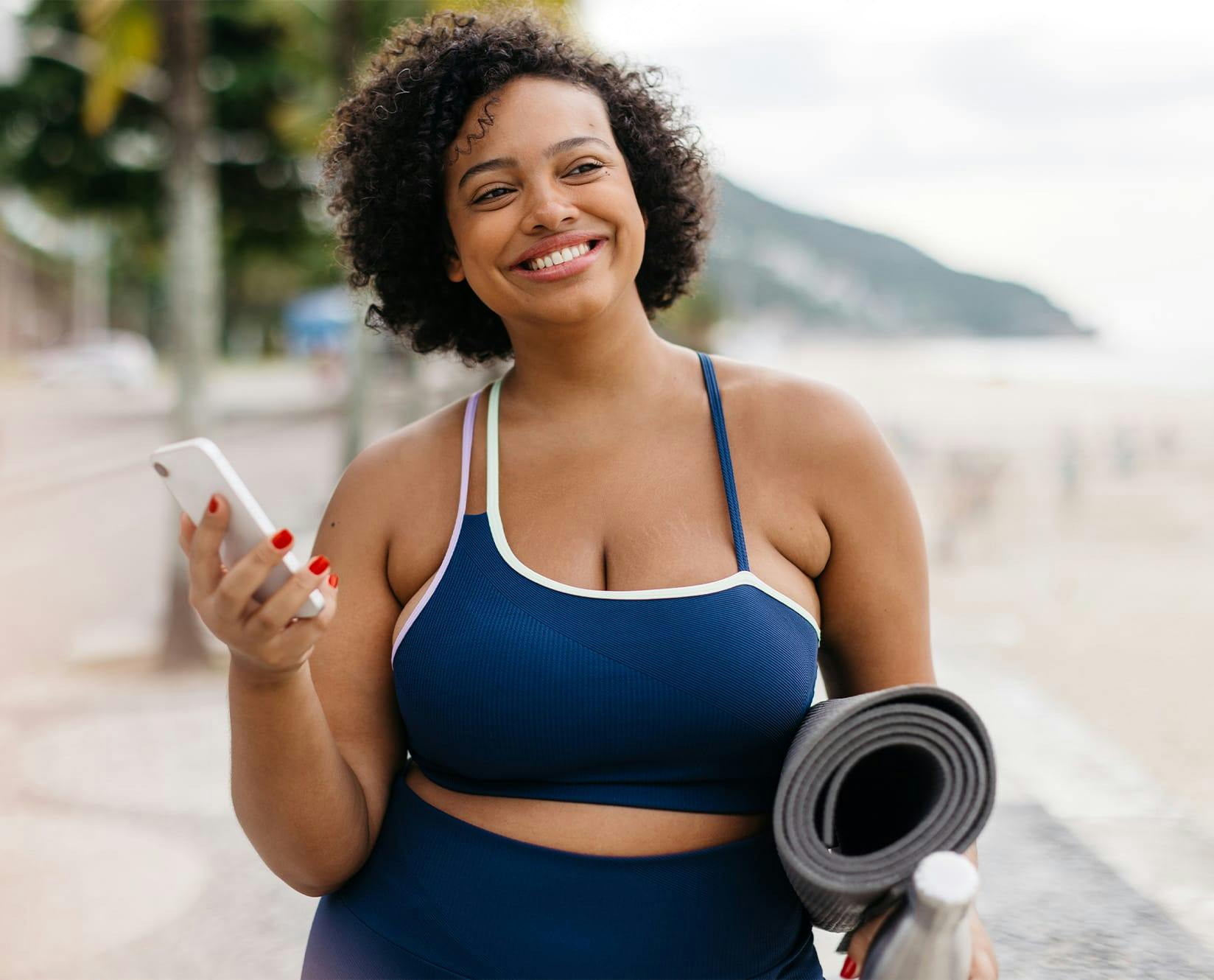 woman with yoga equipment and holding a phone walking on beach
