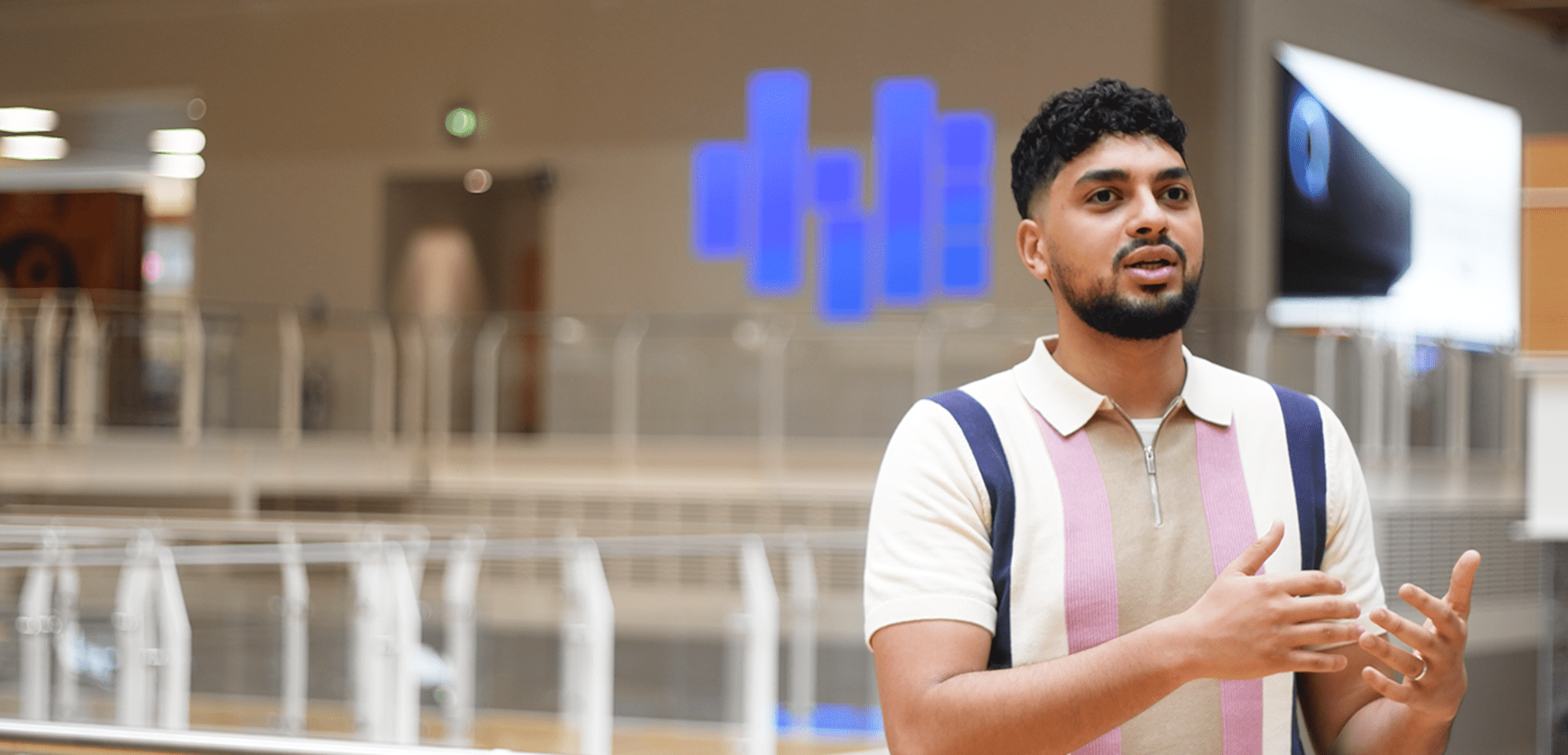 Man speaking with hands raised, wearing a multicoloured polo shirt in a bright, modern indoor setting.