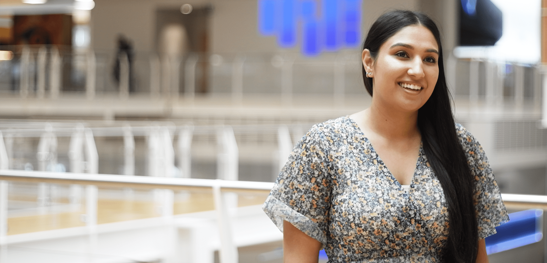 Woman with long dark hair in a floral dress smiling, with bright office space and digital wall lights behind her.