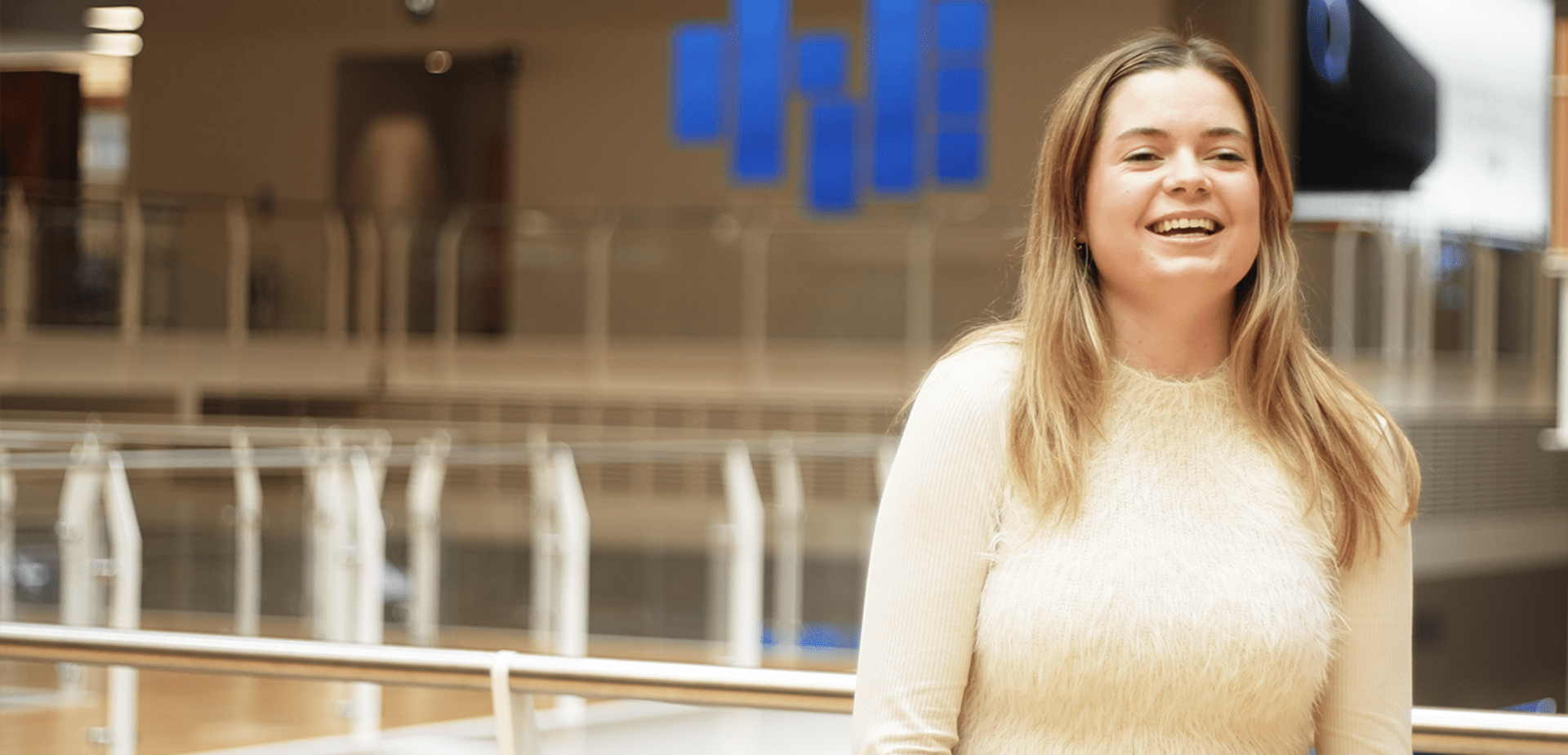 Smiling woman in cream sweater standing in modern office with glass railings and blue logo behind.
