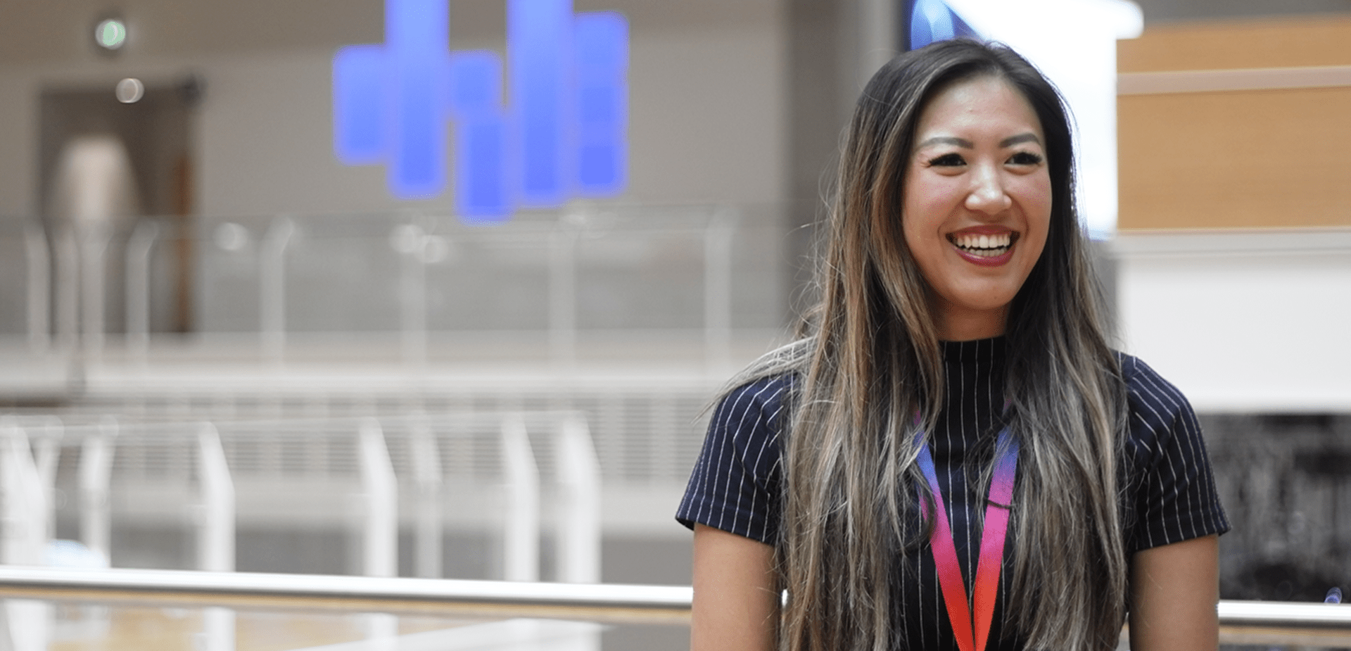 Woman with long hair and striped shirt smiling in a bright, open office atrium.