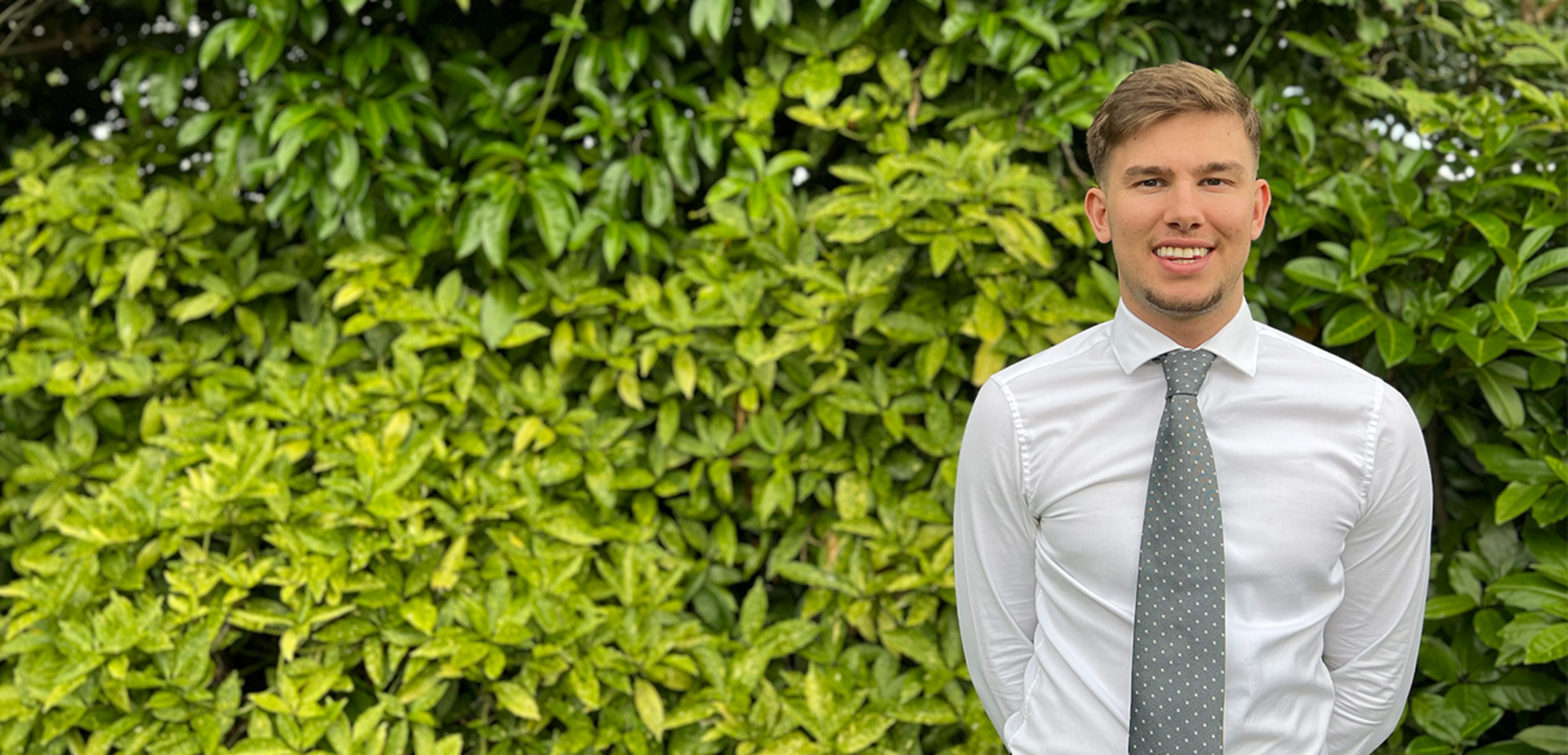 Man in a white shirt and grey tie standing outside in front of a green leafy hedge, smiling with hands behind back.
